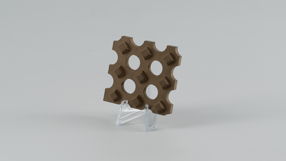 A sample of a geometric design with a metallic sheen, propped up on a small transparent plastic stand. The surface has a complex shape with four equally spaced round through holes and thirteen smaller cubes (protrusions) surrounding them.