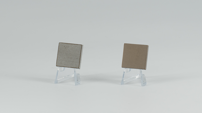 Two square objects propped up on a small transparent plastic stand. The left side is a light gray with a concrete-like texture, while the right side is a subdued beige with a matte metallic sheen.